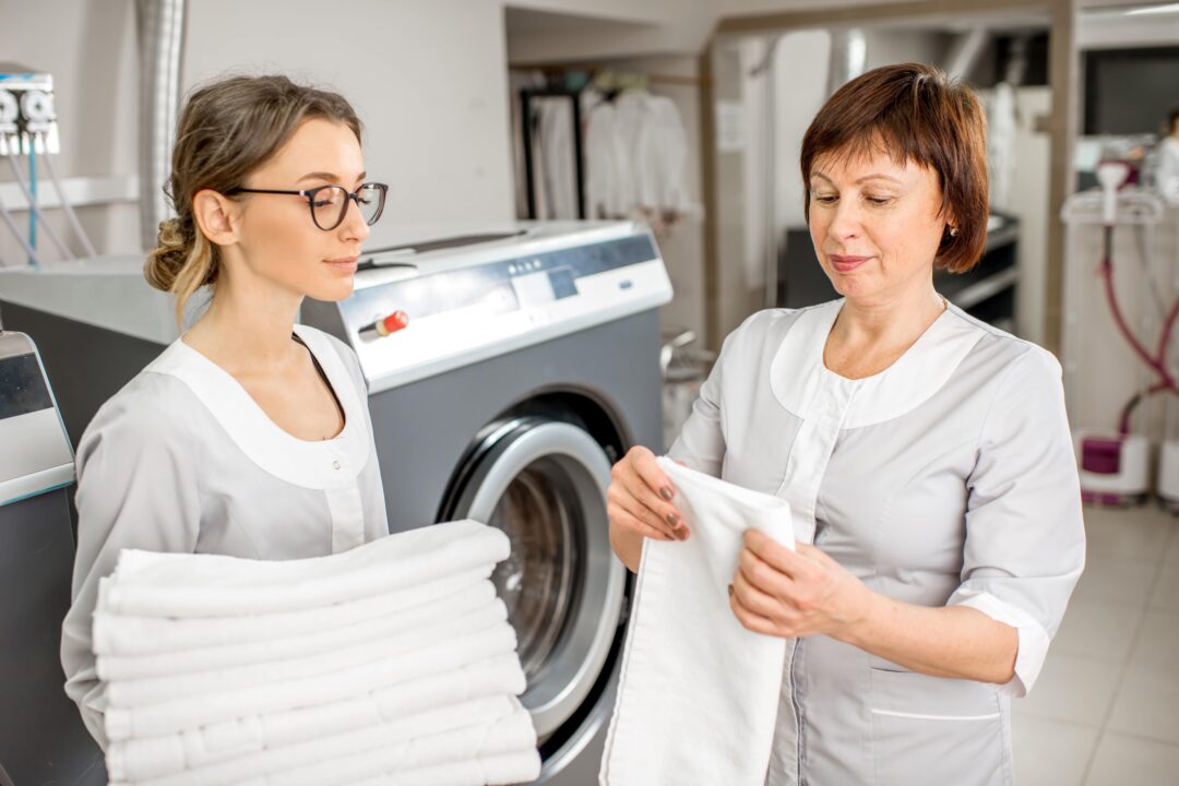 Two hotel staff folding linen sheets in the laundry room.